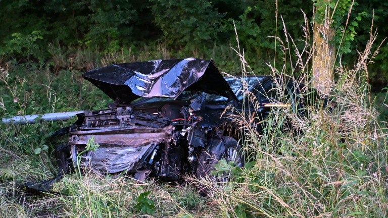 Zwaar beschadigde auto naast de weg in hoog gras, omgeven door bomen.