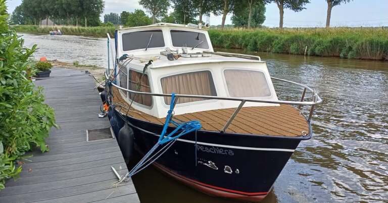 Blauw motorjacht aangemeerd aan een houten steiger bij een kanaal, omgeven door groen en bomen.