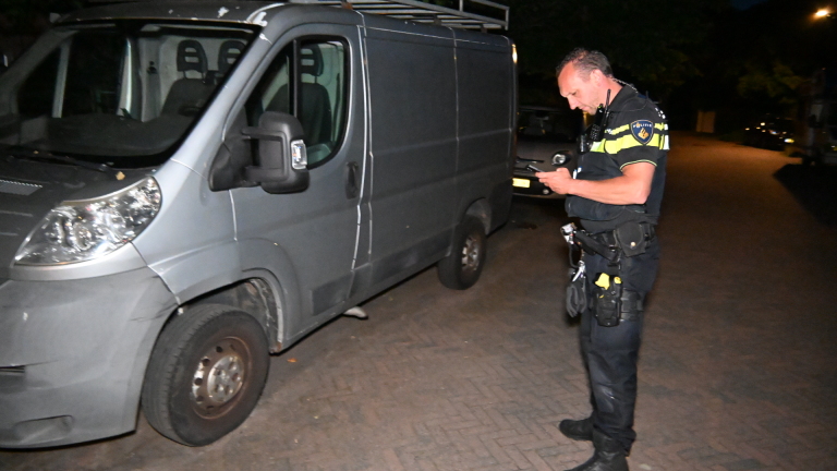 Een politieagent controleert zijn telefoon naast een geparkeerde grijze bestelbus op een straat in het donker.