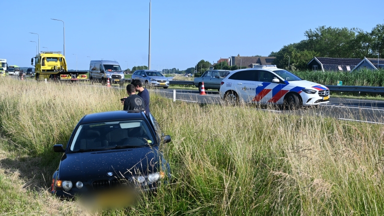 Beschadigde witte auto naast vangrail, met twee mannen in oranje werkkleding dichterbij op de weg.