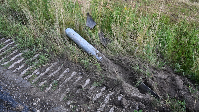 Zwarte auto met beschadigde voorkant staat naast een weg met hoog gras en gele bloemen.