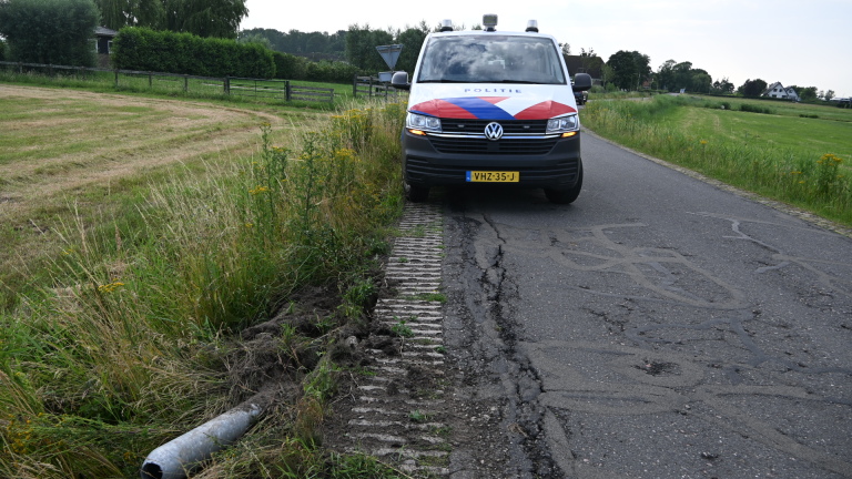 Zwarte auto met beschadigde voorkant staat naast een weg met hoog gras en gele bloemen.