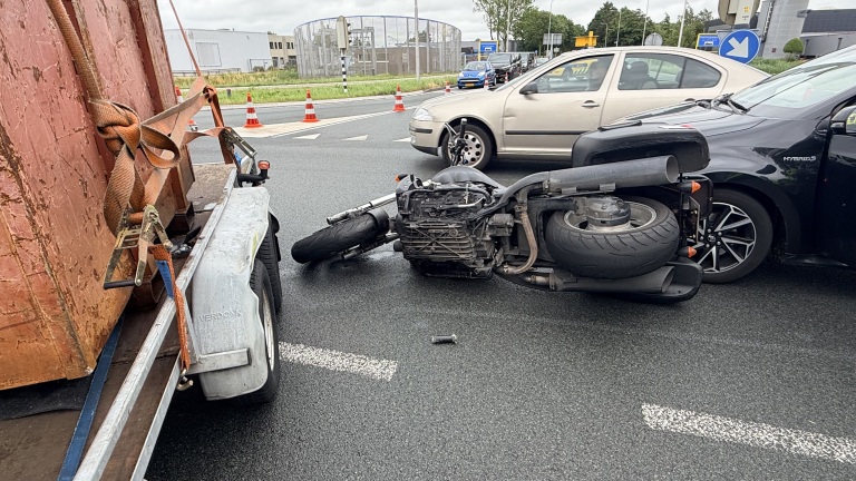 Omgevallen motorfiets op de grond naast een auto en aanhanger op een kruispunt, met verkeer en verkeerskegels op de achtergrond.