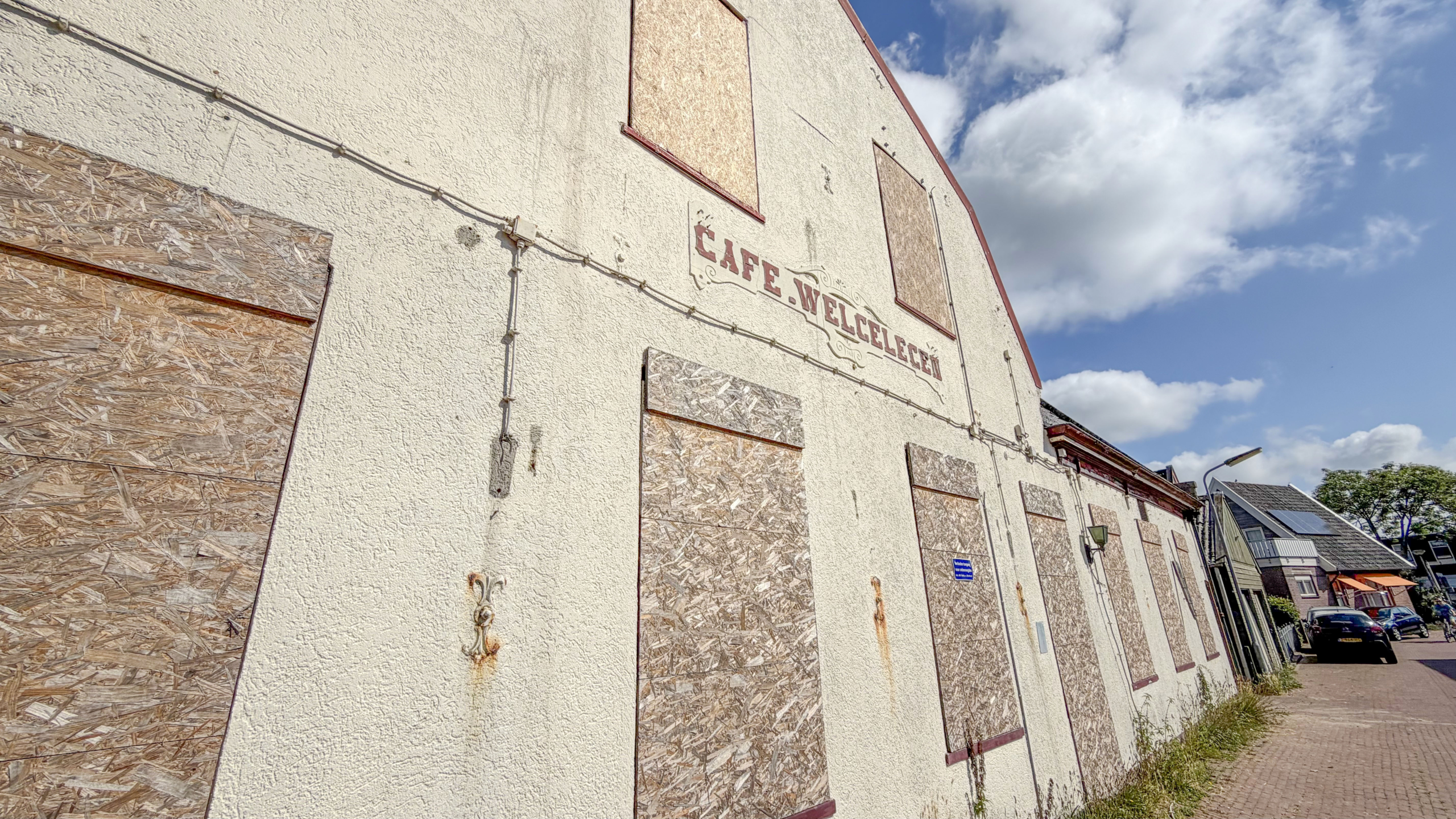 Een verlaten gebouw met dichtgetimmerde ramen, met de tekst "Cafe Welcelecen" op de gevel onder een heldere blauwe hemel.
