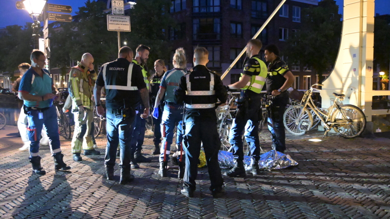 Gele ambulance en hulpverleners bij een verlichte ophaalbrug in de avond, met omstanders op straat