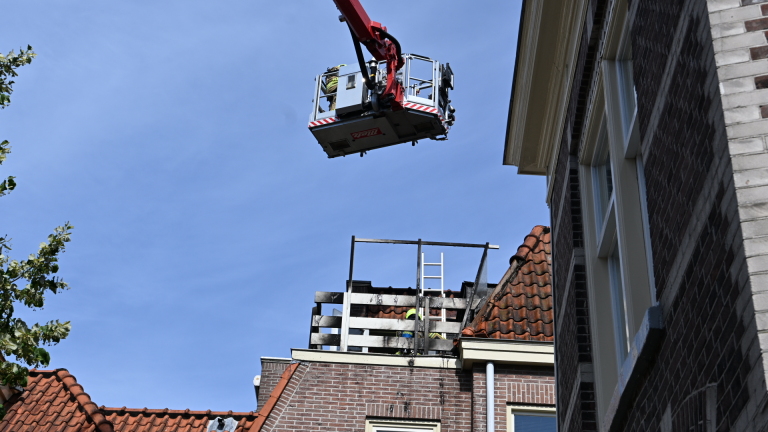 Twee brandweerlieden met gele helmen op een dakbalkon met ladder, bij een beschadigde schoorsteen en dakpannen