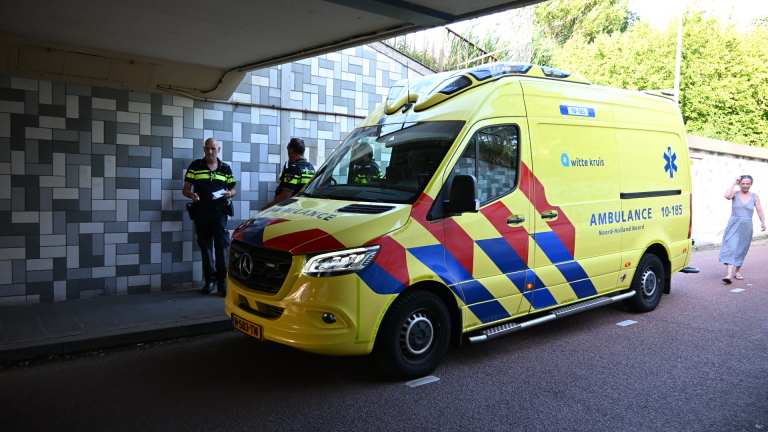 Politieagent loopt in een tunnel langs twee gele ambulances op een fietspad, met daglicht en bomen op de achtergrond