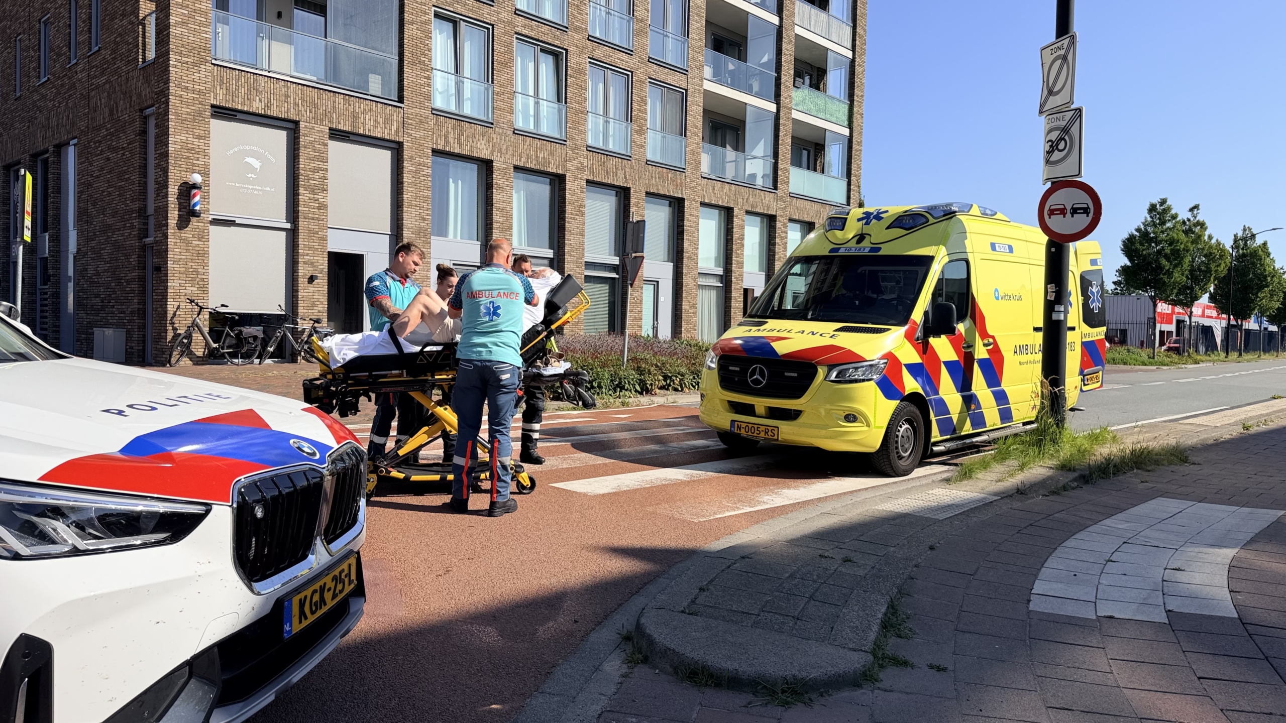 Ambulance en politieauto bij een ongeval, met hulpverleners rond een brancard op een kruispunt voor modern gebouw