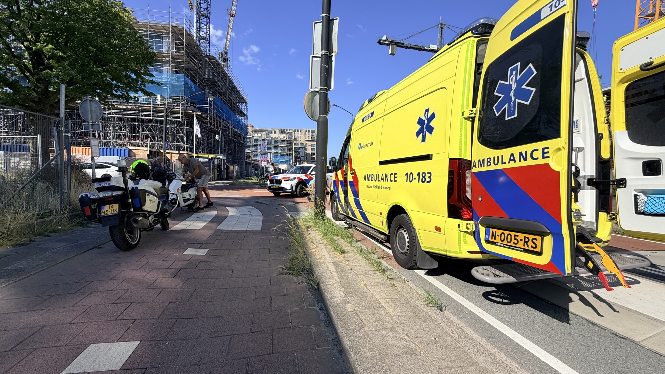Ambulance en politieauto bij een ongeval, met hulpverleners rond een brancard op een kruispunt voor modern gebouw
