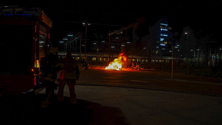 Brand bij fietsenstalling naast een station, met brandweerwagen en brandweerlieden terwijl rook en vlammen opstijgen