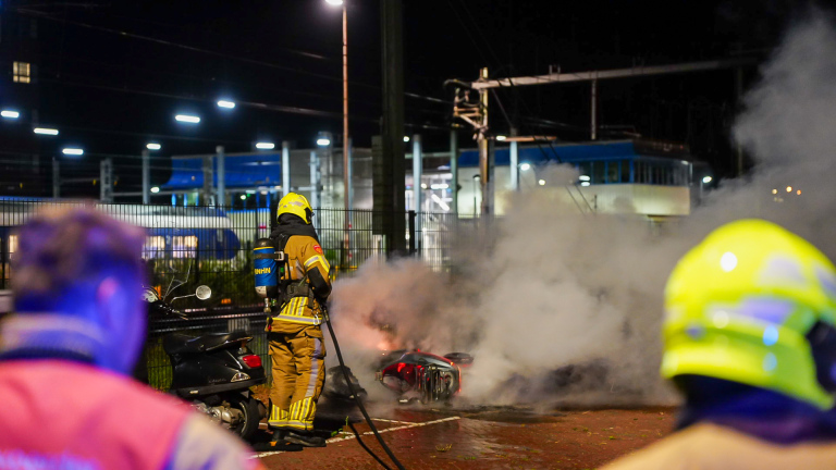 Brand bij fietsenstalling naast een station, met brandweerwagen en brandweerlieden terwijl rook en vlammen opstijgen