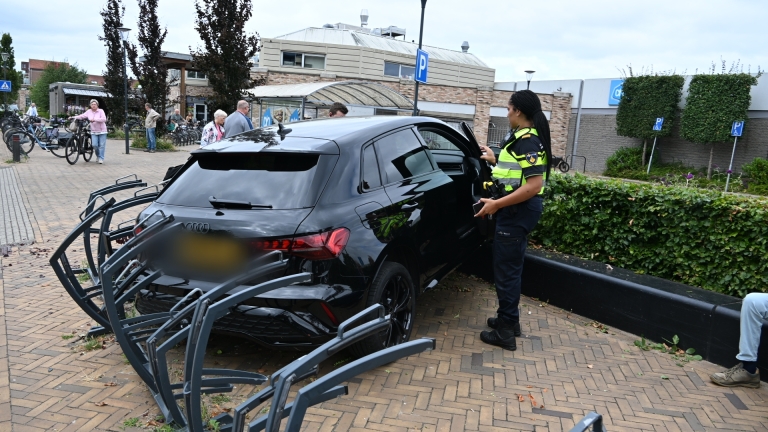 Zwarte auto op stoep tegen fietsenrek gereden, met afgebroken boomtakken ervoor bij een supermarkt