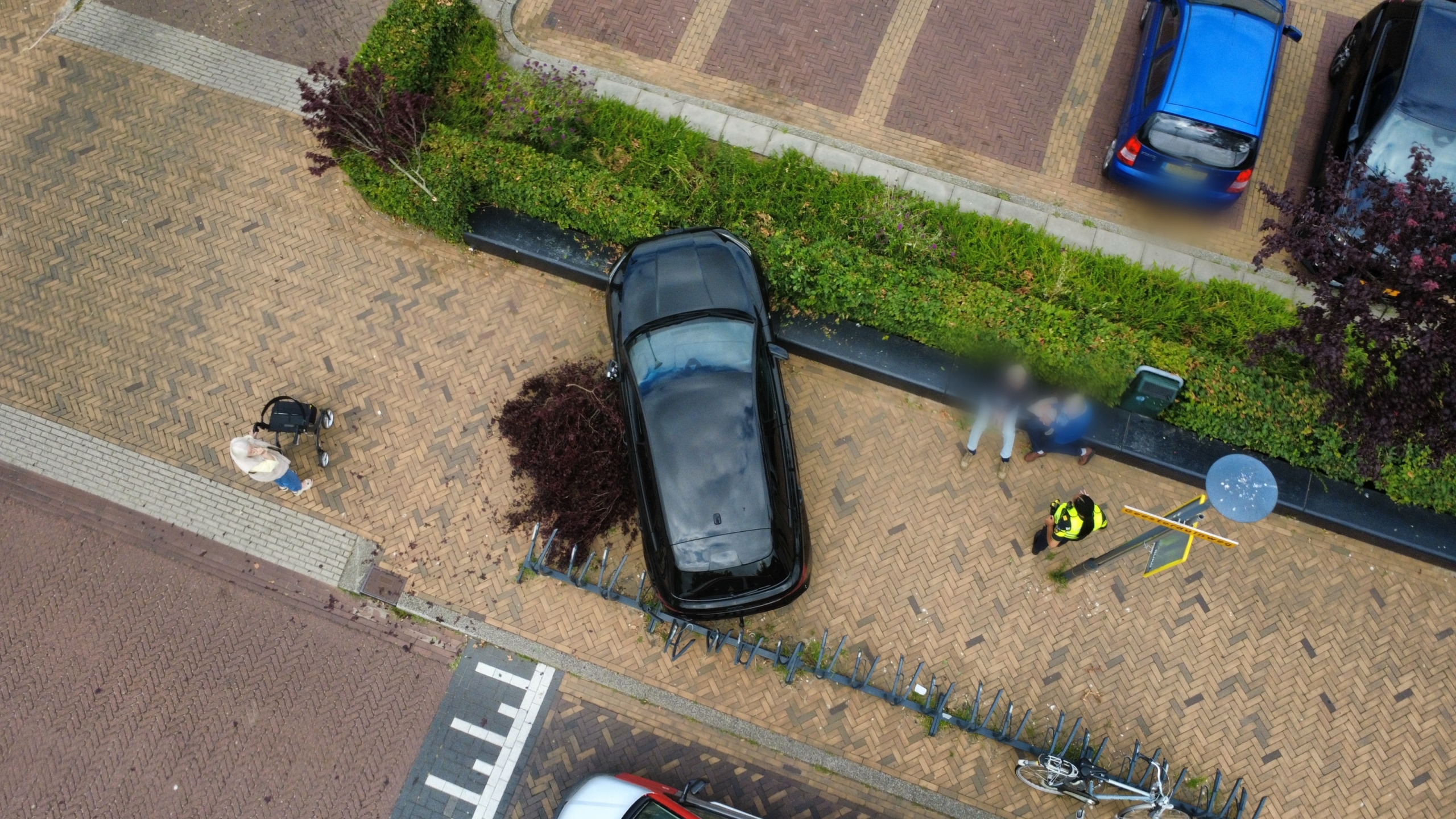 Zwarte auto op stoep tegen fietsenrek gereden, met afgebroken boomtakken ervoor bij een supermarkt