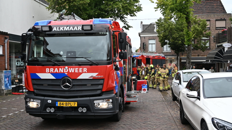 Brandweerauto met open achterklep en brandweerlieden op een straat bij bakstenen panden en winkel Kaldi