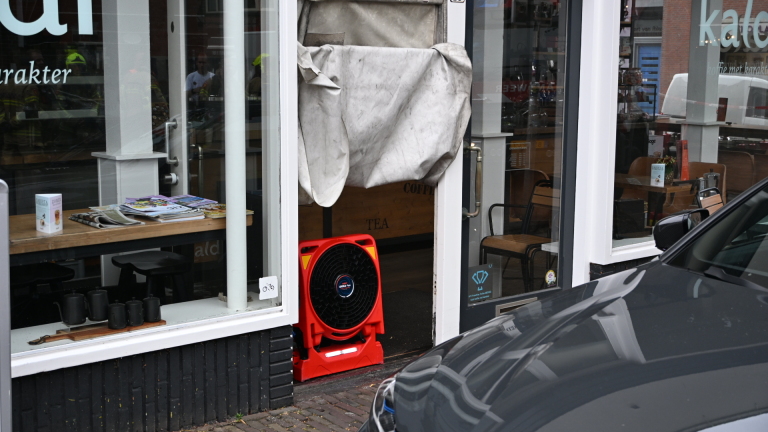 Brandweerauto met open achterklep en brandweerlieden op een straat bij bakstenen panden en winkel Kaldi