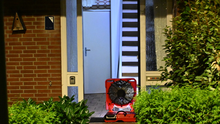 Dikke rookwolken stijgen ’s nachts op achter huizen, met fel verlichte groene struiken op de voorgrond
