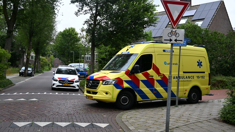 Ambulance van Witte Kruis staat bij fietspad; hulpverlener naast een fiets en afzetkegel, voetgangers op de achtergrond