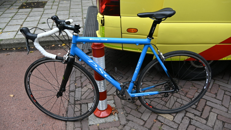 Ambulance van Witte Kruis staat bij fietspad; hulpverlener naast een fiets en afzetkegel, voetgangers op de achtergrond