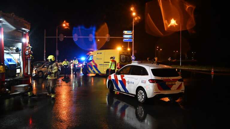 Zwaar beschadigde auto op nat wegdek na aanrijding, met ambulance en hulpverleners op de achtergrond