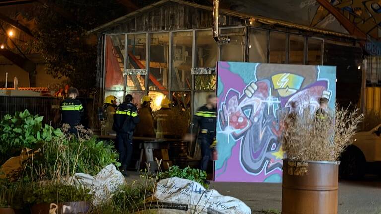Brandweerlieden staan ’s avonds bij een deels uitgebrande loods met gebroken ruiten en een groot graffiti-paneel ervoor