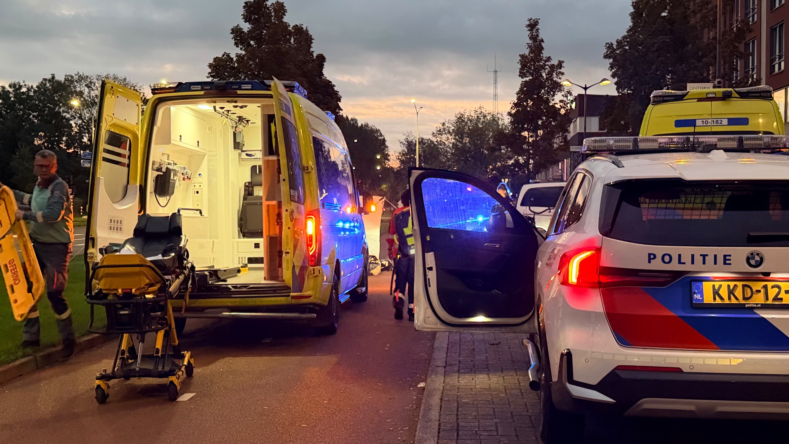 Twee ambulances met hulpverleners die iemand behandelen bij een wit afschermdoek langs de weg in de schemering