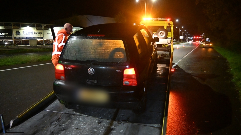Zwarte auto deels in een sloot beland langs een grasberm, met modder op de carrosserie en koplampen aan